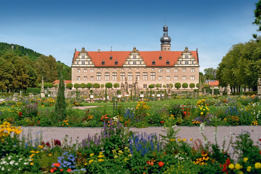Ein barockes Schloss mit rotem Ziegeldach und Turm vor bunter Blumenpracht