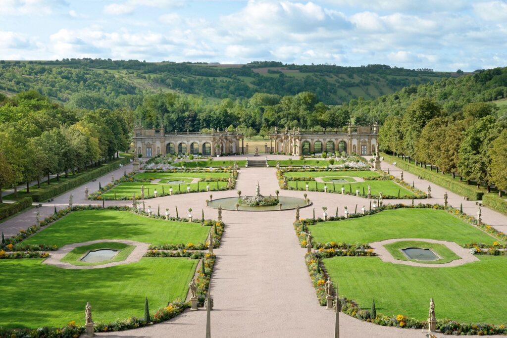 Weitläufiger Garten mit wegen und Grünflächen, im Hintergrund das lang gezogene Gebäude einer Orangerie