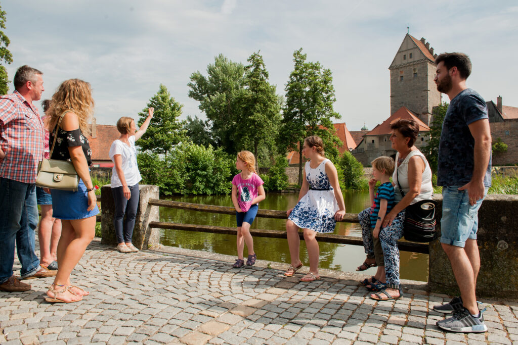 Eine Gruppe von Erwachsenen und Kindern bekommt von einer Gästeführerin etwas erklärt. Im Hintergrund sind ein Turm und Bäume zu sehen.