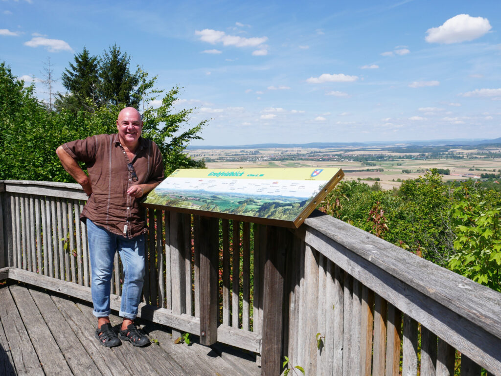 Ein Mann steht an einem Aussichtspunkt, im Hintergrund der Blick über Felder und Bäume.