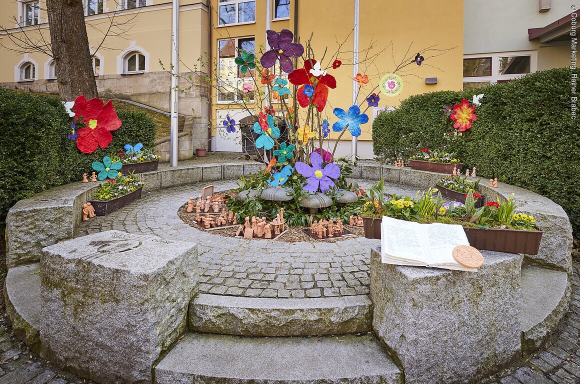 Ein kleiner Brunnen in Mitten einer Steinfläche, bunt geschmückt mit Blumen