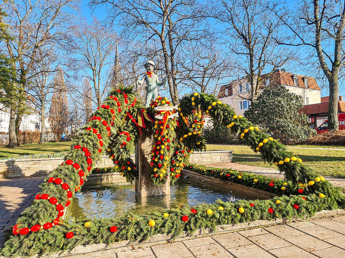 Ein bunt geschmückter Osterbrunnen mit einer kleinen Figur darauf, dahinter ein grüner Park