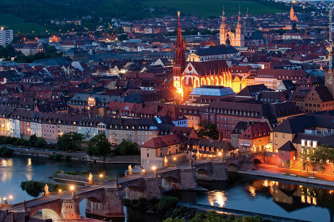 Panoramablick über eine abendliche Stadt an einem Fluss. Zu sehen sind Brücken, Häuser, Kirchen und Türme