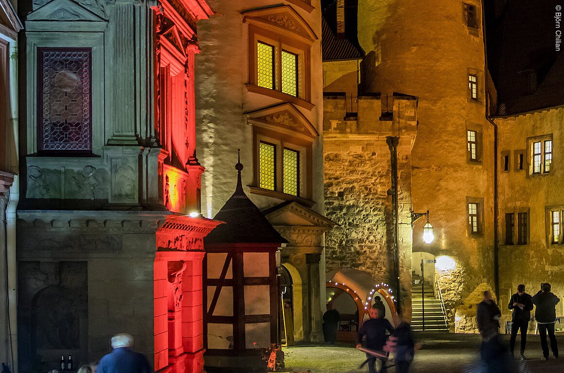 Historische Gebäude bei Nacht, beleuchtet in verschiedenen Farben, mit einem Turm im Hintergrund.