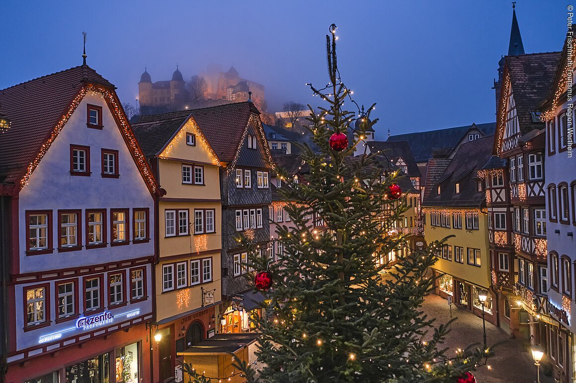 Weihnachtlicher Marktplatz und Burg im Nebel Beleuchtete Fachwerkhäuser und ein geschmückter Weihnachtsbaum in einer abendlichen Stadtansicht.