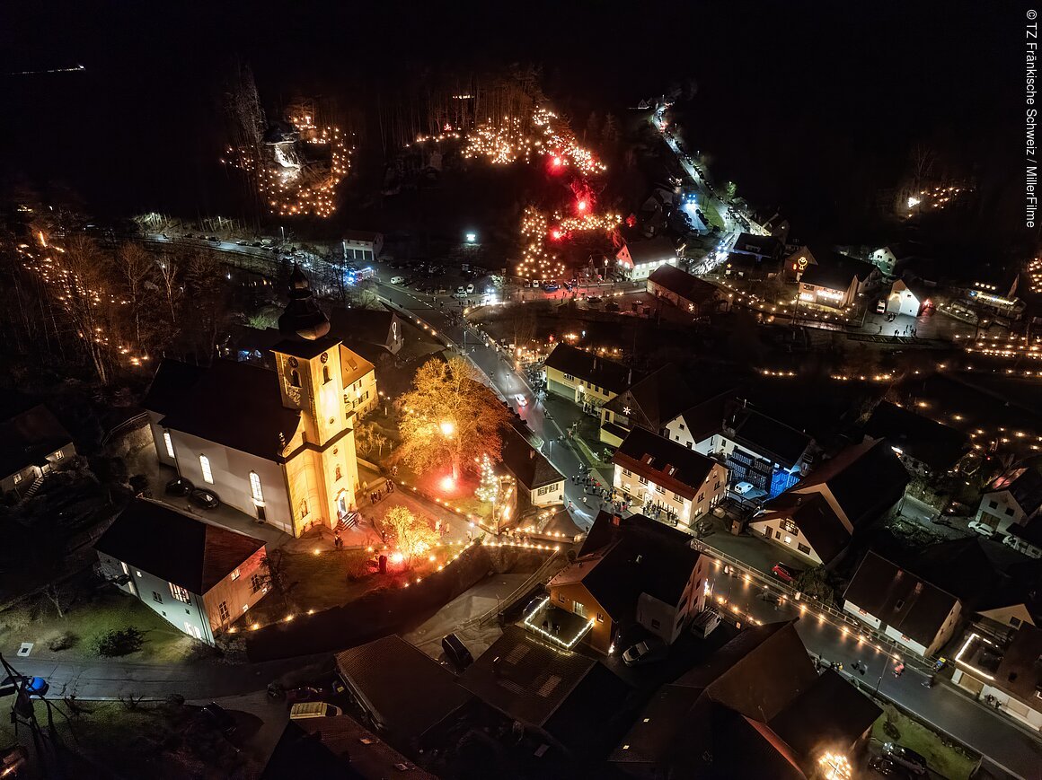 Luftaufnahme eines fränkischen Dorfes bei Nacht, beleuchtet von zahlreichen Lichtern und einer erleuchteten Kirche im Vordergrund.
