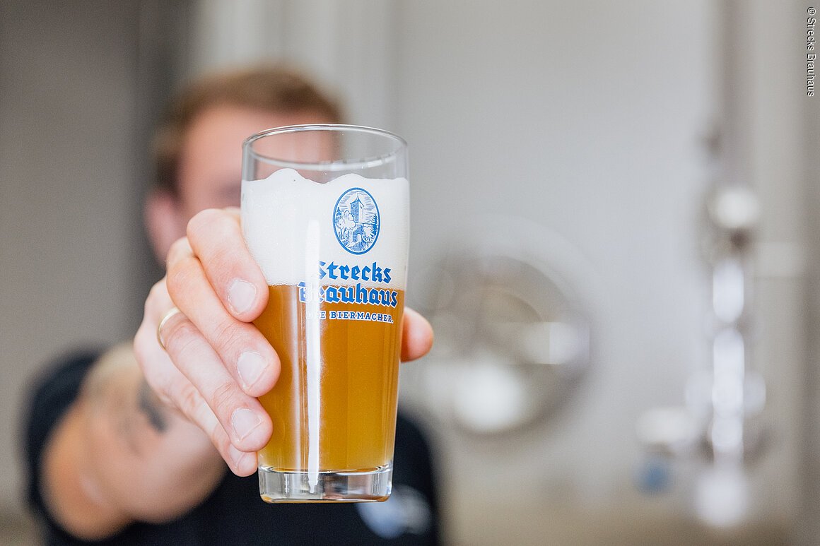 Eine Person hält ein Glas Bier mit Schaumkrone, darauf steht "Strecks Brauhaus".