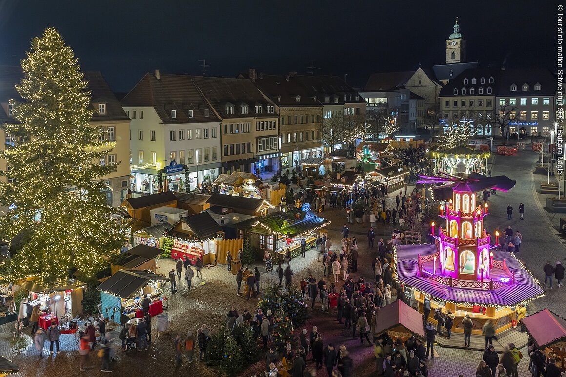 Weihnachtsmarkt Schweinfurt bei Nacht mit beleuchtetem Baum und Ständen, Menschenmenge auf dem Platz.