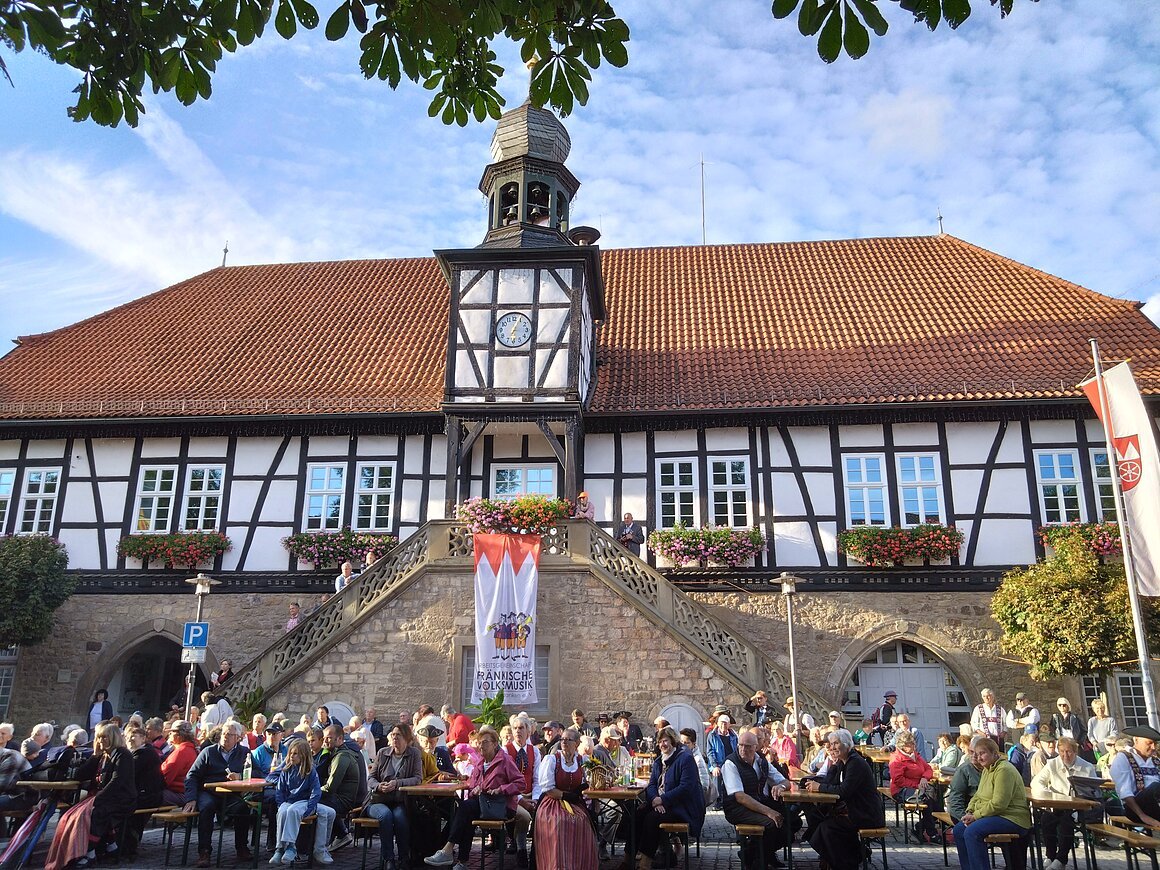 Rathaus (Ostheim v.d.Rhön, Rhön) Fachwerkgebäude mit rotem Dach, Menschen sitzen an Tischen davor, blauer Himmel im Hintergrund.