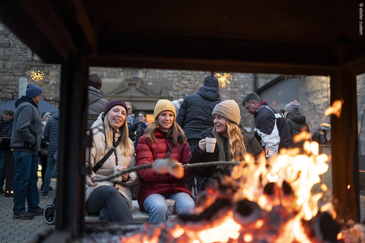 Drei Personen sitzen vor einem Feuer, halten Tassen und rösten Marshmallows. Im Hintergrund sind weitere Menschen.