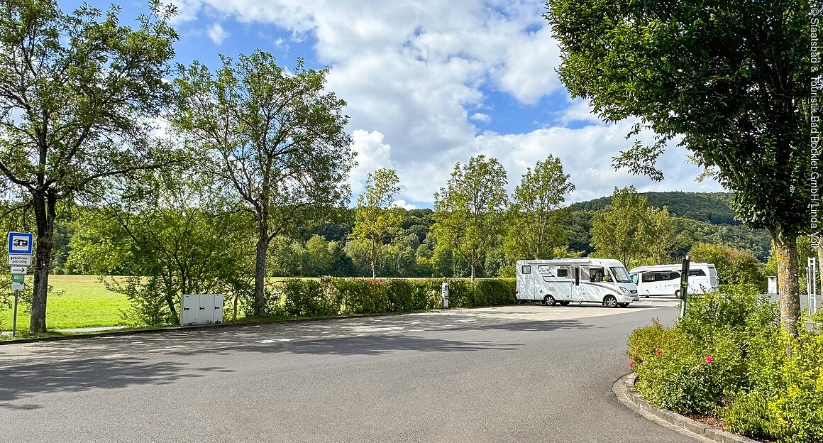 Wohnmobilstellplatz (Bad Bocklet, Rhön) Wohnmobile parken auf einem Rastplatz mit Bäumen und grüner Landschaft im Hintergrund.