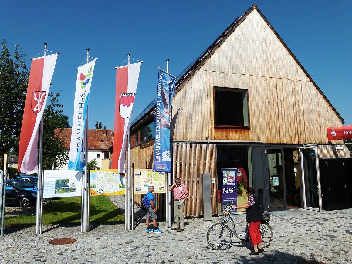 Altmühlsee Informationszentrum (Muhr a. See, Fränkisches Seenland) Modernes Gebäude mit Holzverkleidung, davor mehrere Fahnen und Informationstafeln. Eine Person mit Fahrrad steht vor dem Eingang.