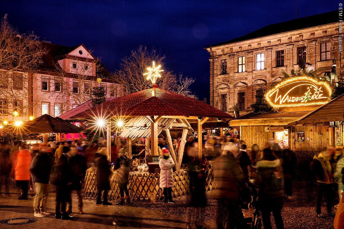 Menschenmenge auf Erlanger Weihnachtsmarkt bei Nacht, umgeben von beleuchteten Holzhütten und Dekorationen.