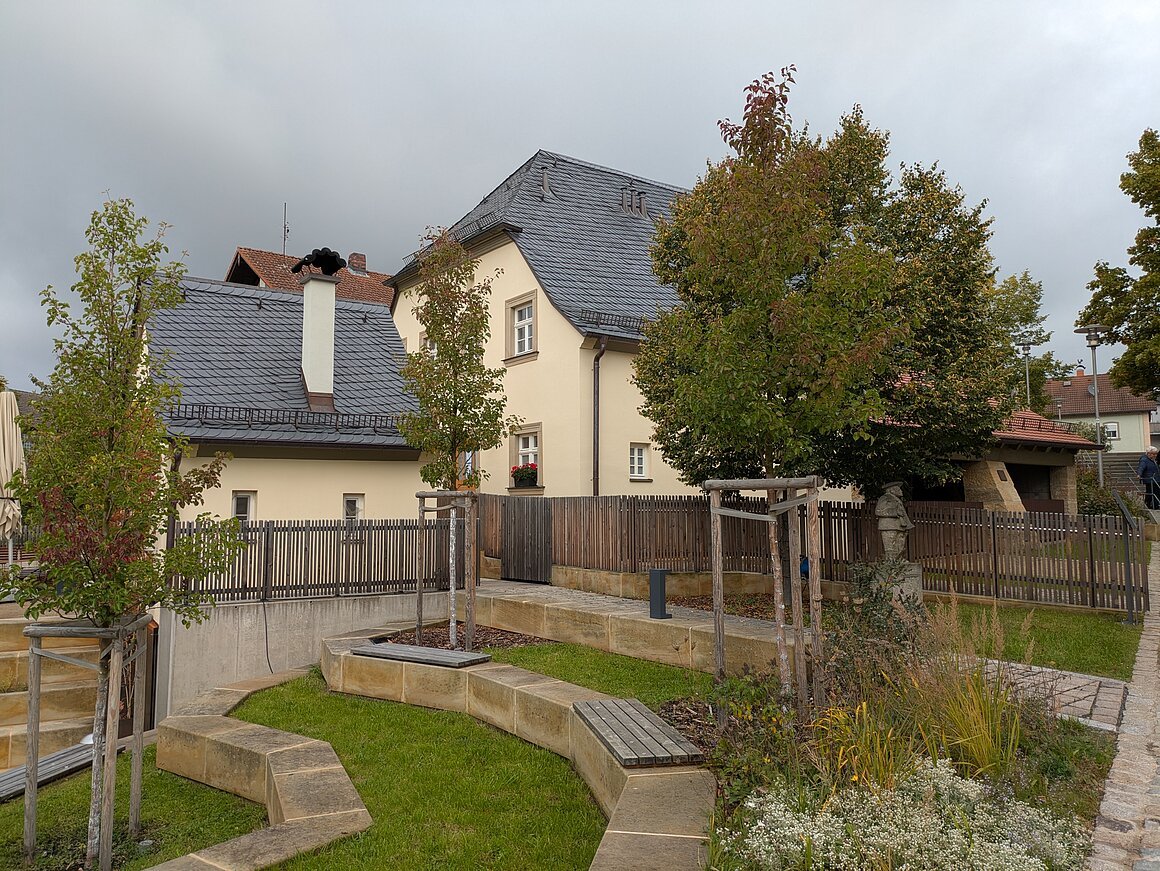 Haus mit Schieferdach, Garten mit Bäumen und Bänken, Holzzaun im Hintergrund.