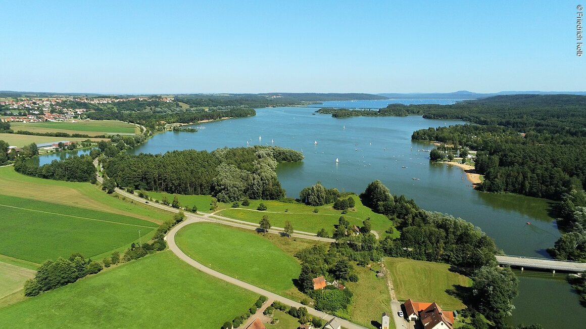 Luftaufnahme eines Sees mit Segelbooten, umgeben von Wäldern und Wiesen. Im Hintergrund sind Hügel sichtbar.