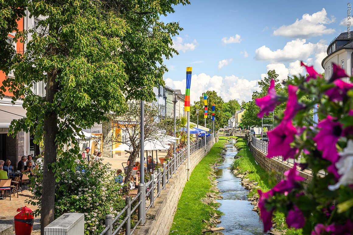 Stadtansicht (Münchberg, Fichtelgebirge) Stadtansicht mit kleinem Fluss, bunten Fahnen, Bäumen und Menschen in einem Café.
