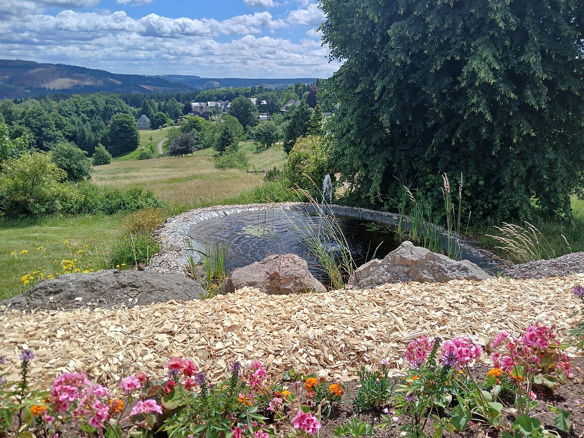 Rathausvorplatz (Masserberg, Coburg.Rennsteig) Garten mit Teich, Blumen und Holzschnitzelweg, im Hintergrund eine hügelige Landschaft mit Bäumen und Häusern.