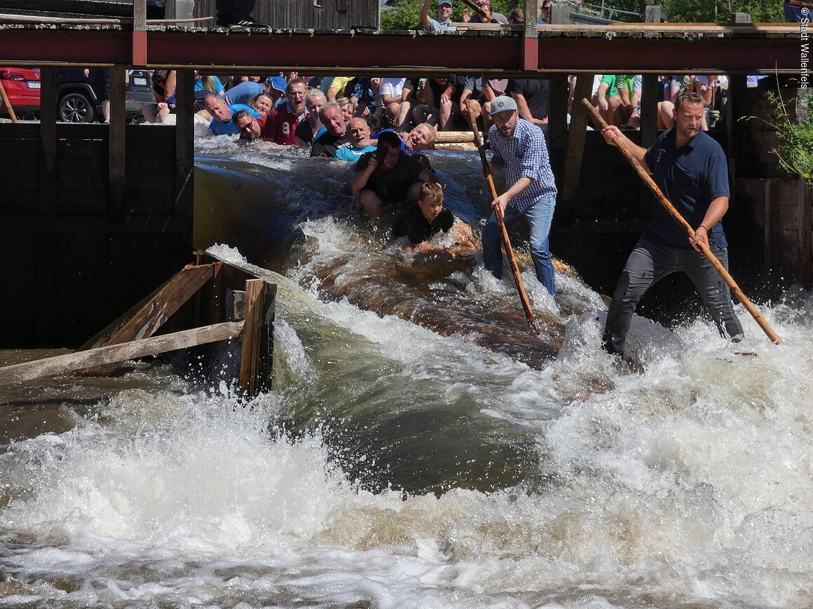 Floßfahrt auf der Wilden Rodach (Wallenfels, Frankenwald) Gruppe von Menschen auf einem Floß in einem Fluss, umgeben von spritzendem Wasser.