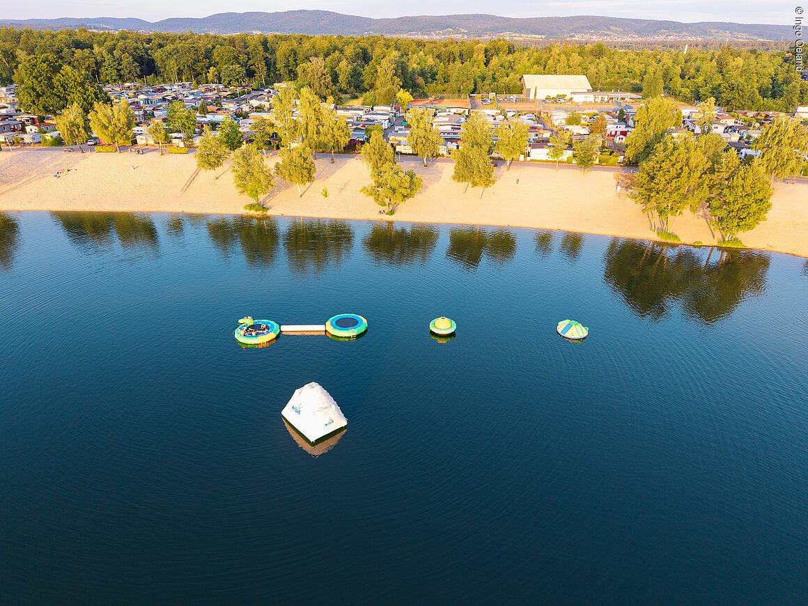 Luftaufnahme eines Sees mit schwimmenden Spielgeräten, Sandstrand und Campingplatz im Hintergrund.