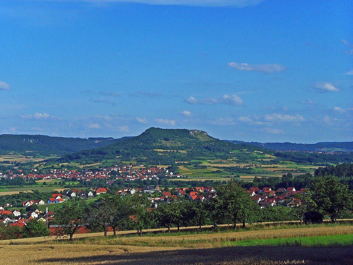 Walberla (Kirchehrenbach, Fränkische Schweiz) Landschaft mit Hügeln, Feldern und einem Dorf mit roten Dächern im Vordergrund.