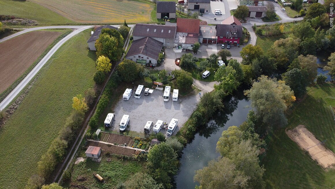 Luftaufnahme eines Campingplatzes mit Wohnmobilen, umgeben von Bäumen, einem Fluss und landwirtschaftlichen Gebäuden.