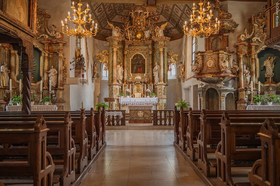 Innenraum einer barocken Kirche mit reich verziertem Altar, Holzbänken und Kronleuchtern.