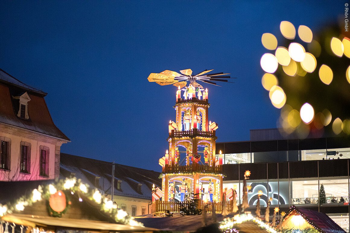 Beleuchtete Weihnachtspyramide auf einem Weihnachtsmarkt bei Nacht, mit unscharfen Lichtern im Vordergrund.