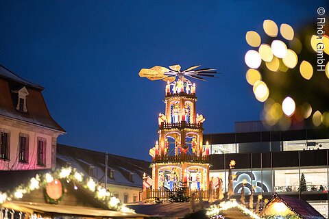 Beleuchtete Weihnachtspyramide auf einem Weihnachtsmarkt bei Nacht, mit unscharfen Lichtern im Vordergrund.