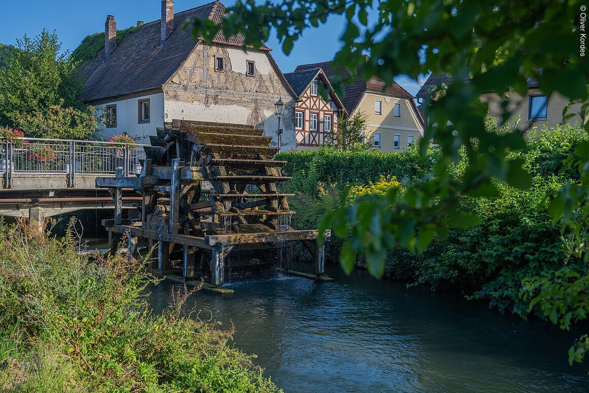 Altes Wasserrad an einem Fluss, umgeben von traditionellen Fachwerkhäusern und üppigem Grün.