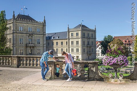 Eine bunt gekleidete Familie ist vor einem Schloss, welches aus mehreren Bauten besteht, zu sehen. Im Hintergrund ist ein Schlossgarten erkennbar.