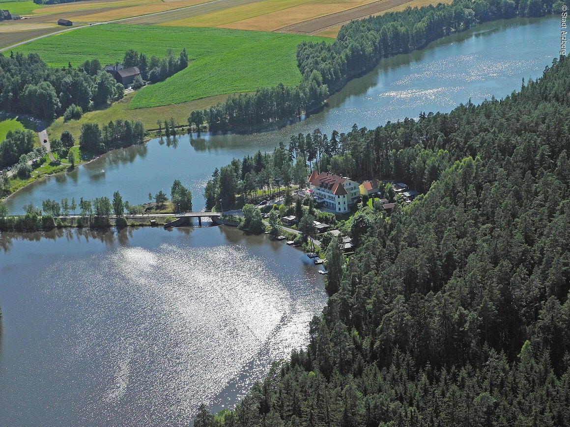 Luftaufnahme eines Sees mit angrenzendem Wald und Gebäuden am Ufer. Eine Brücke überquert den See.