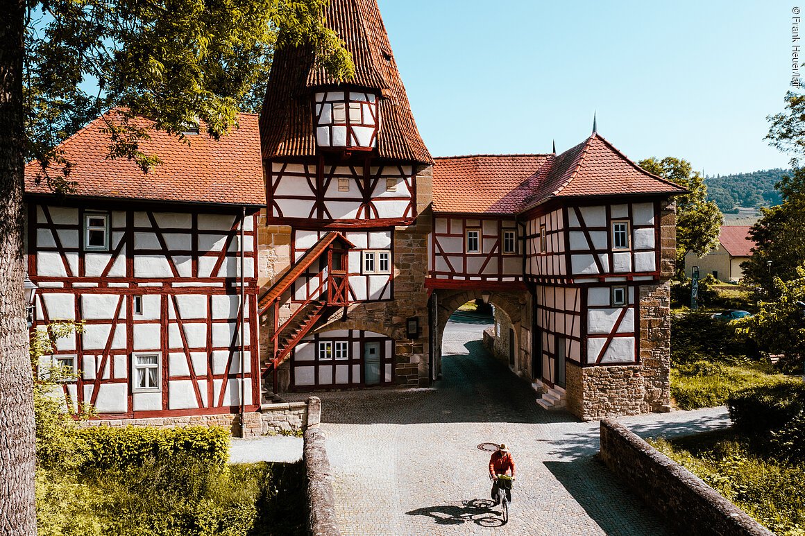 Rödelseer Tor (Iphofen, Fränkisches Weinland) Fachwerkgebäude mit Torbogen und rotem Dach, Radfahrer auf gepflastertem Weg. Bäume im Hintergrund.