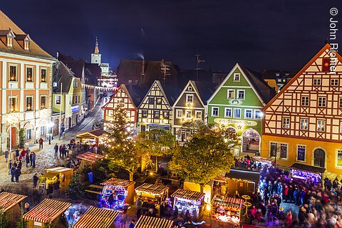 Belebter Weihnachtsmarkt mit Fachwerkhäusern im Hintergrund, beleuchtet bei Nacht.