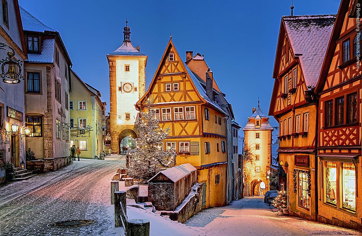 Plönlein im Winter (Rothenburg o.d.Tauber, Romantisches Franken) Verschneite Altstadt mit Fachwerkhäusern und beleuchteten Straßenlaternen bei Dämmerung. Ein Turm im Hintergrund.