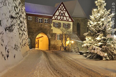 Verschneite Straße führt durch ein beleuchtetes Tor in einem Fachwerkgebäude. Rechts steht ein beleuchteter Weihnachtsbaum.