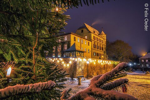 Verschneiter Weihnachtsbaum mit Lichtern im Vordergrund, beleuchtetes Gebäude im Hintergrund.