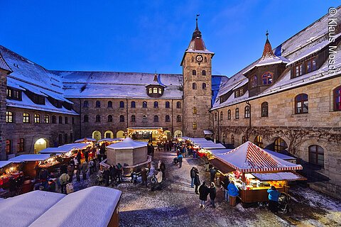 Weihnachtsmarkt in einem schneebedeckten Innenhof mit beleuchteten Ständen und einem Uhrturm.