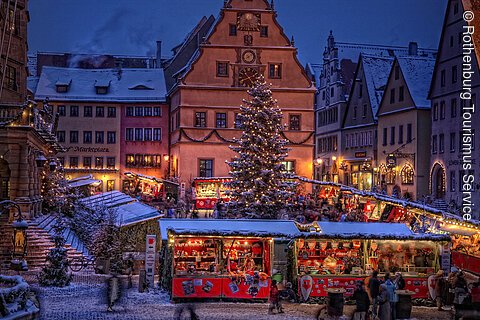 Weihnachtsmarkt mit geschmücktem Baum, beleuchteten Ständen und schneebedeckten Gebäuden in der Dämmerung.