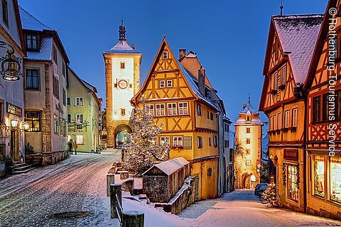 Verschneite Altstadt mit Fachwerkhäusern und beleuchteten Straßenlaternen bei Dämmerung. Ein Turm im Hintergrund.