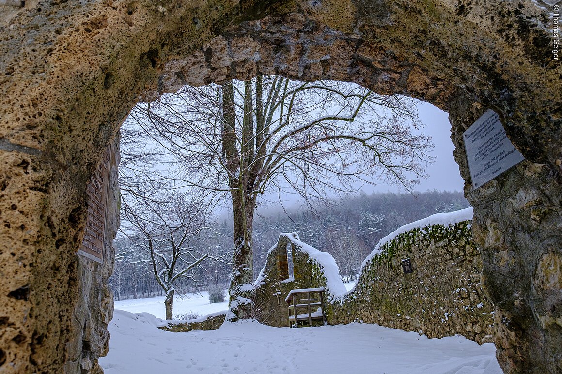 Steinbogen mit Blick auf verschneite Landschaft, Bäume und eine Steinmauer im Hintergrund.