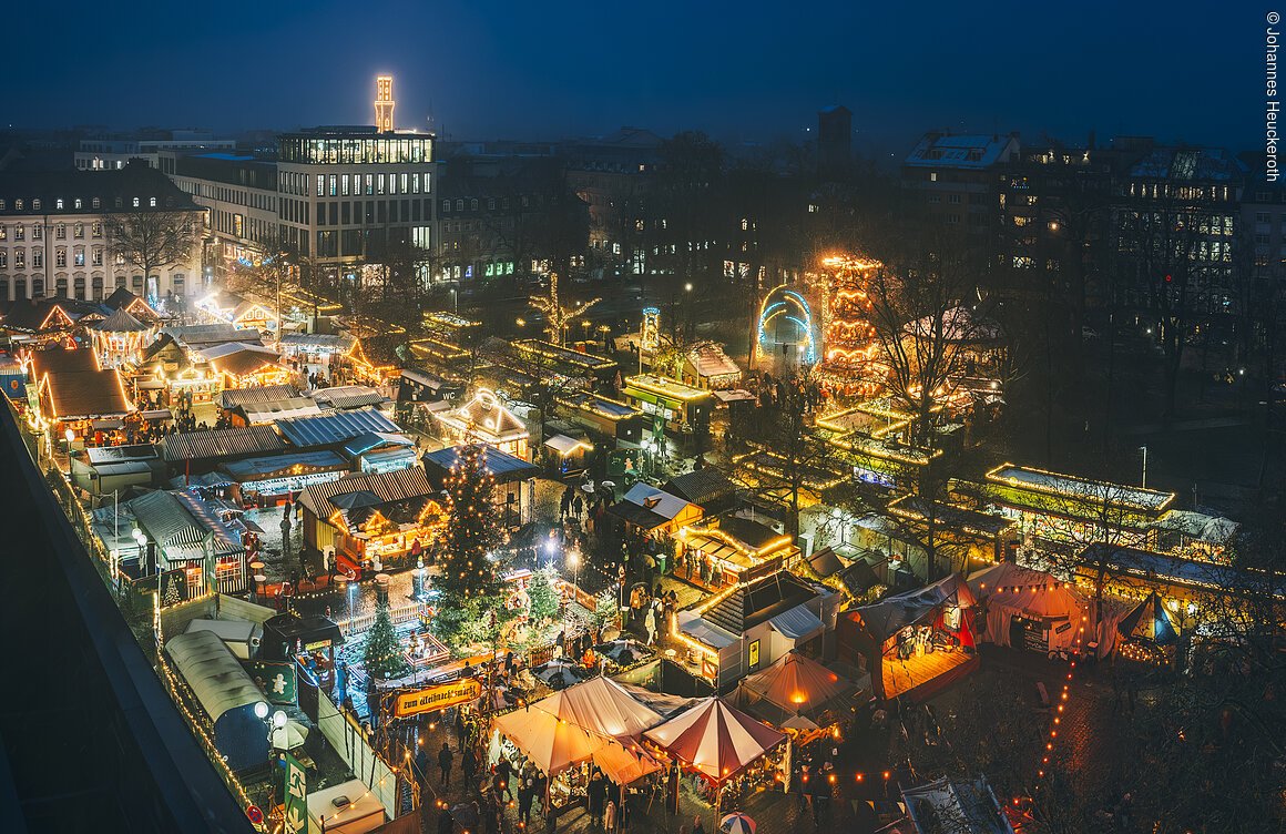 Luftaufnahme eines Weihnachtsmarktes bei Nacht mit beleuchteten Ständen und einem großen Weihnachtsbaum.