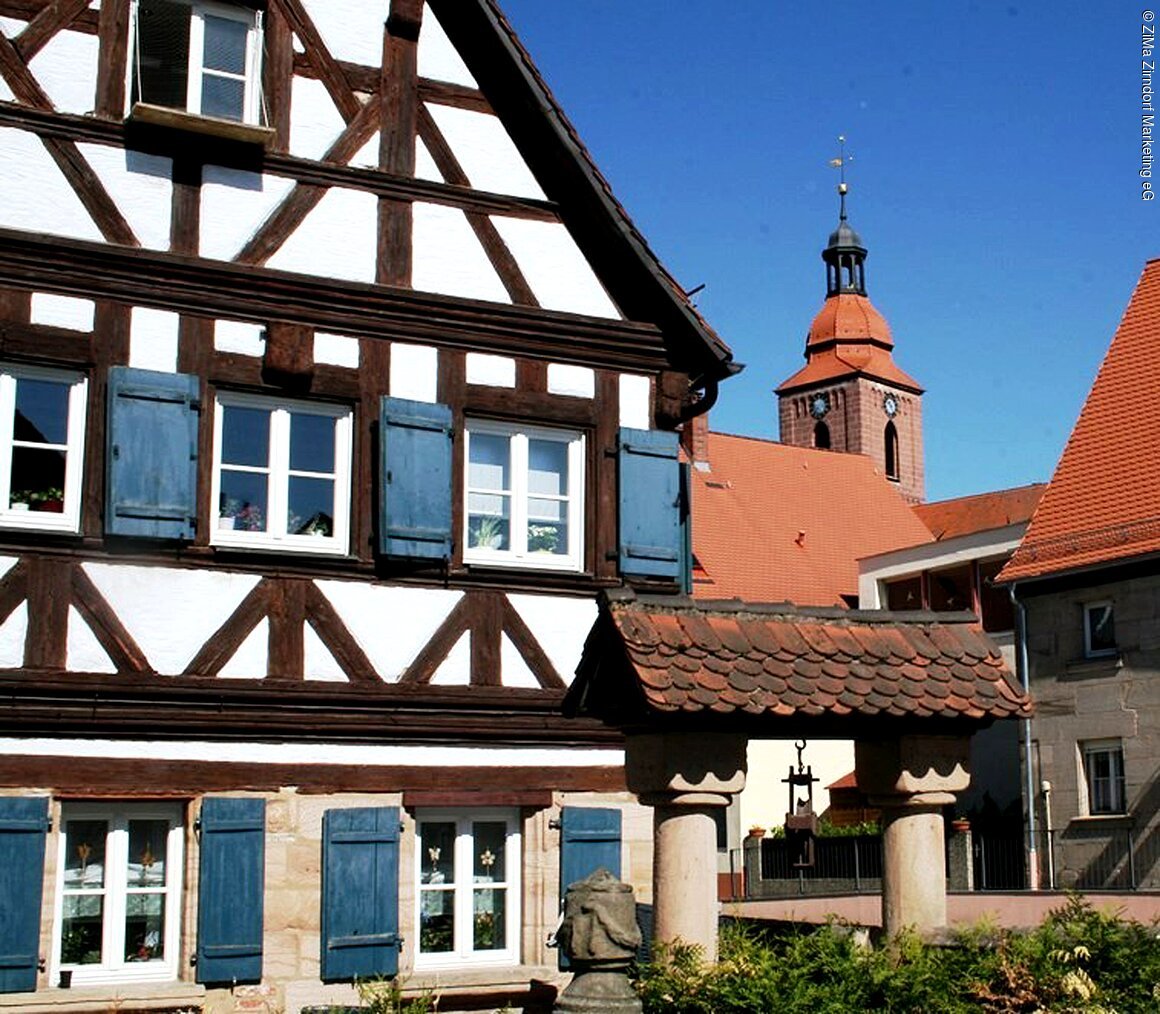 Fachwerkhaus mit blauen Fensterläden, im Hintergrund ein Kirchturm mit Uhr und rotem Dach.