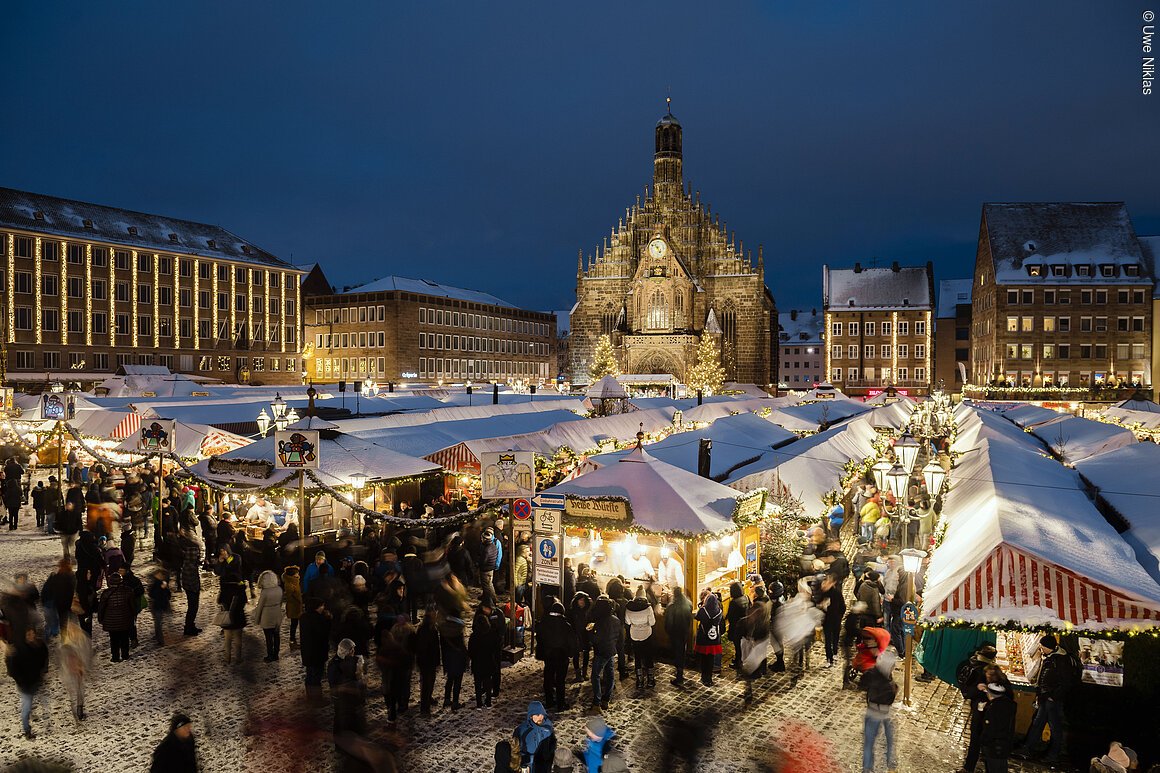 Weihnachtsmarkt mit beleuchteten Ständen und Menschenmenge bei Nacht, im Hintergrund eine Kirche.