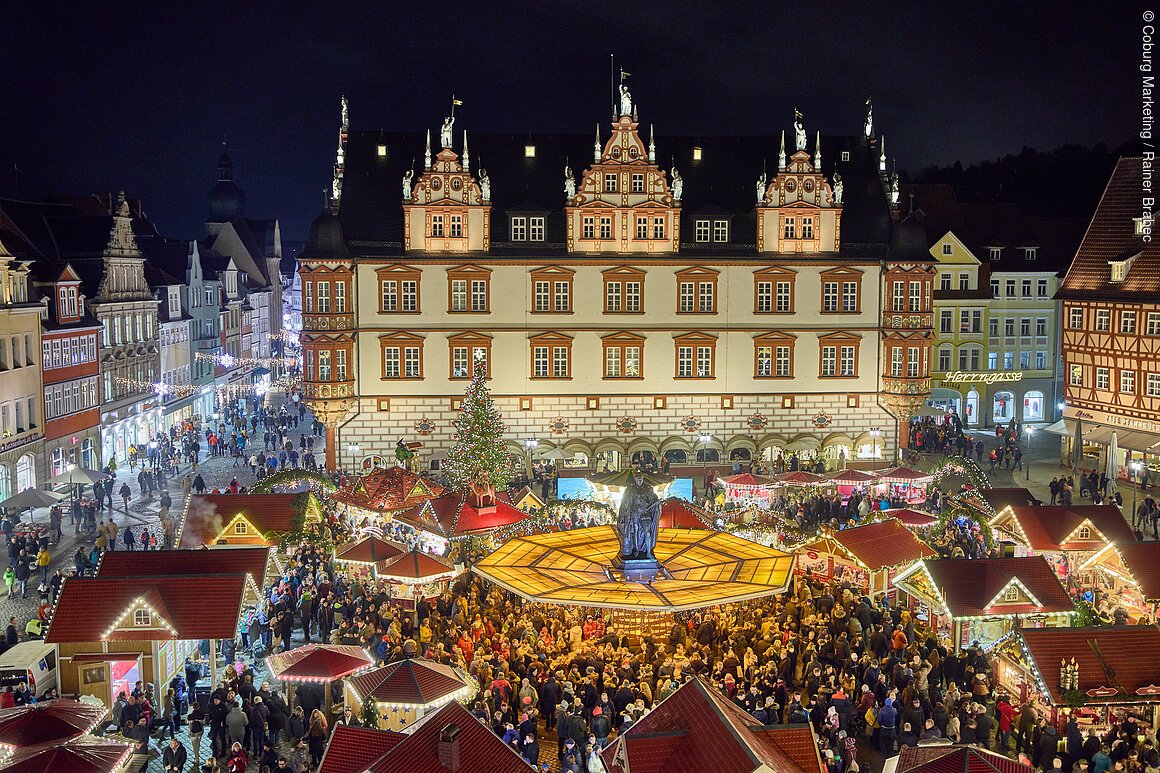 Weihnachtsmarkt bei Nacht mit beleuchteten Ständen, einem großen Weihnachtsbaum und einem historischen Gebäude im Hintergrund.
