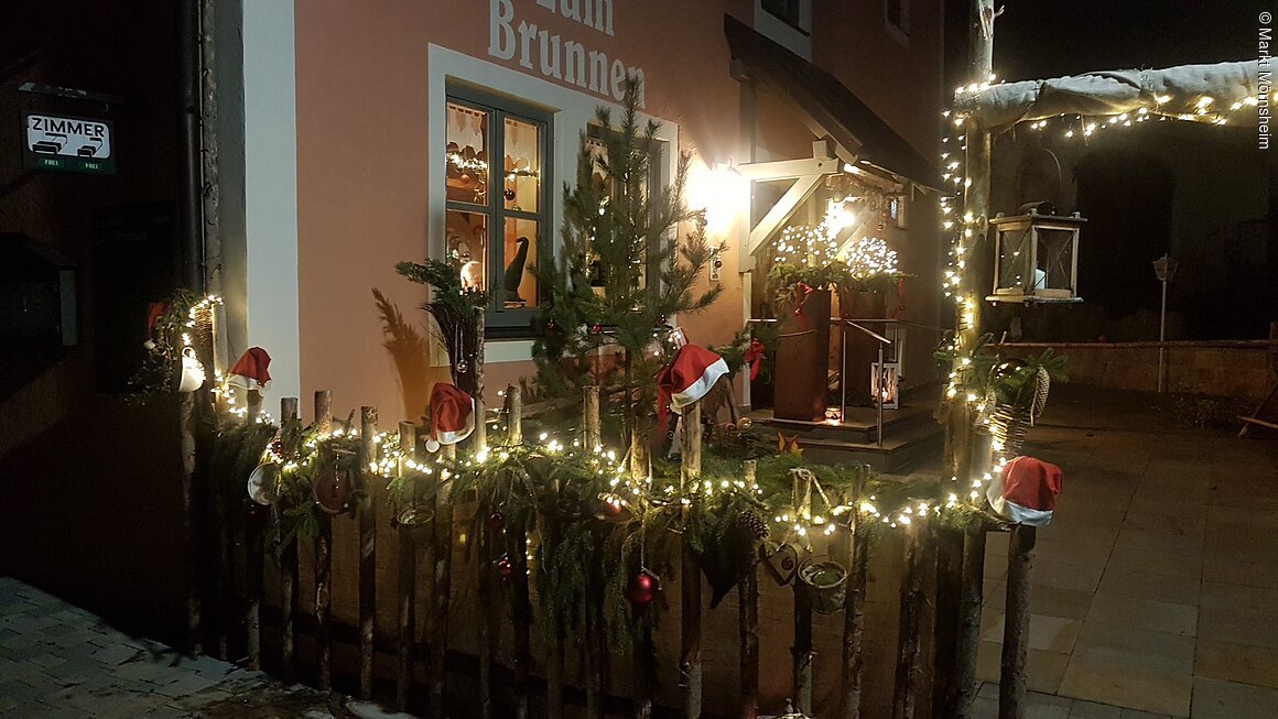 Weihnachtlich dekorierter Eingang mit Lichterketten, Tannenzweigen und Nikolausmützen an einem Zaun.