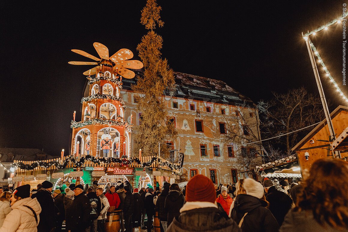Menschenmenge auf einem Weihnachtsmarkt bei Nacht, mit beleuchteter Weihnachtspyramide und dekoriertem Gebäude im Hintergrund.