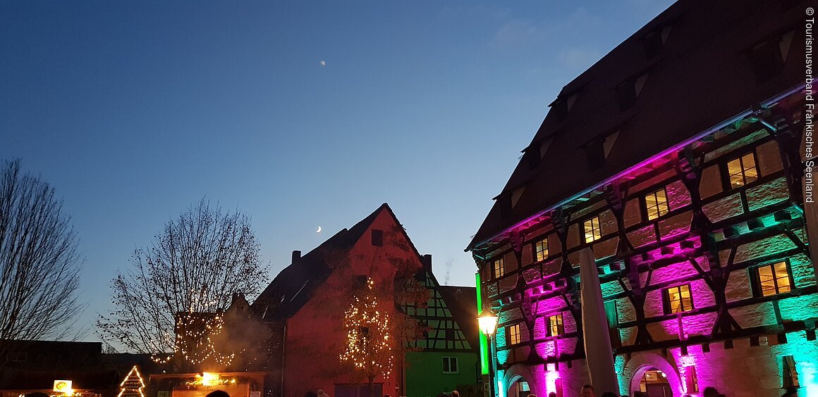 Fachwerkhäuser bei Dämmerung, beleuchtet in bunten Farben. Im Vordergrund Weihnachtsdekoration und Lichter.