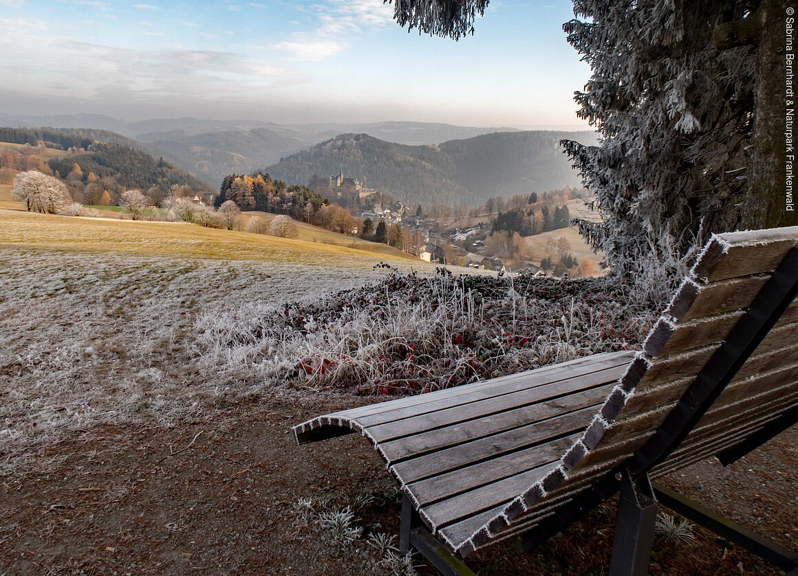Holzbank mit Raureif, im Vordergrund, blickt auf eine frostige Landschaft mit Hügeln, Bäumen und einem Dorf im Hintergrund.