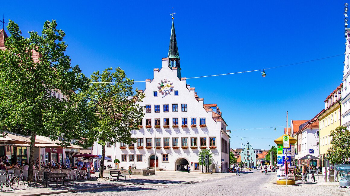 Ein historisches Gebäude mit Giebel und Turm steht auf einem Platz, umgeben von Bäumen und Cafés.
