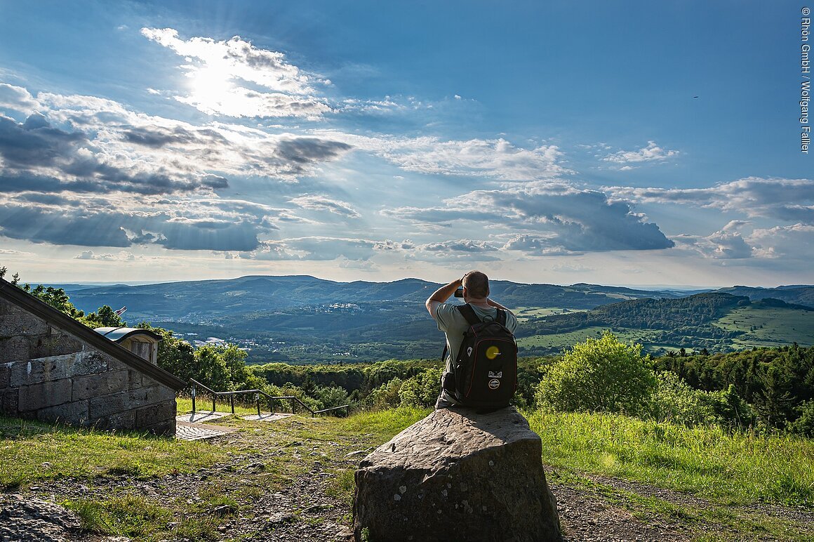 Person sitzt auf einem Felsen und betrachtet eine hügelige Landschaft unter einem bewölkten Himmel.